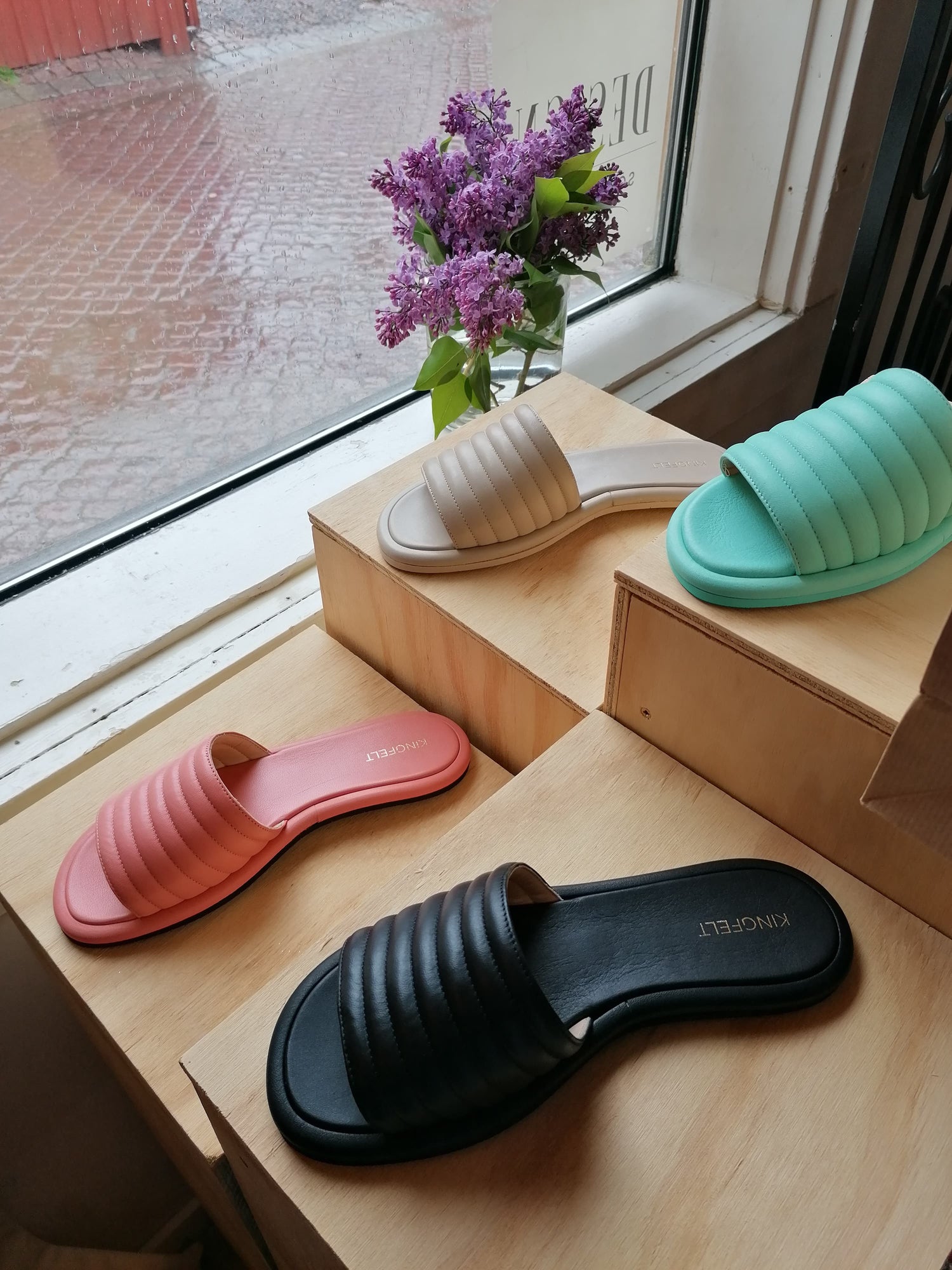 Skinnsandaler i fyra olika färger i skyltfönster hos Design Söderköping. Hållbara sandaler tillverkade i Italien. Svarta, beige, korallrosa och mintgröna sandaler på pidestaler vid fönster.