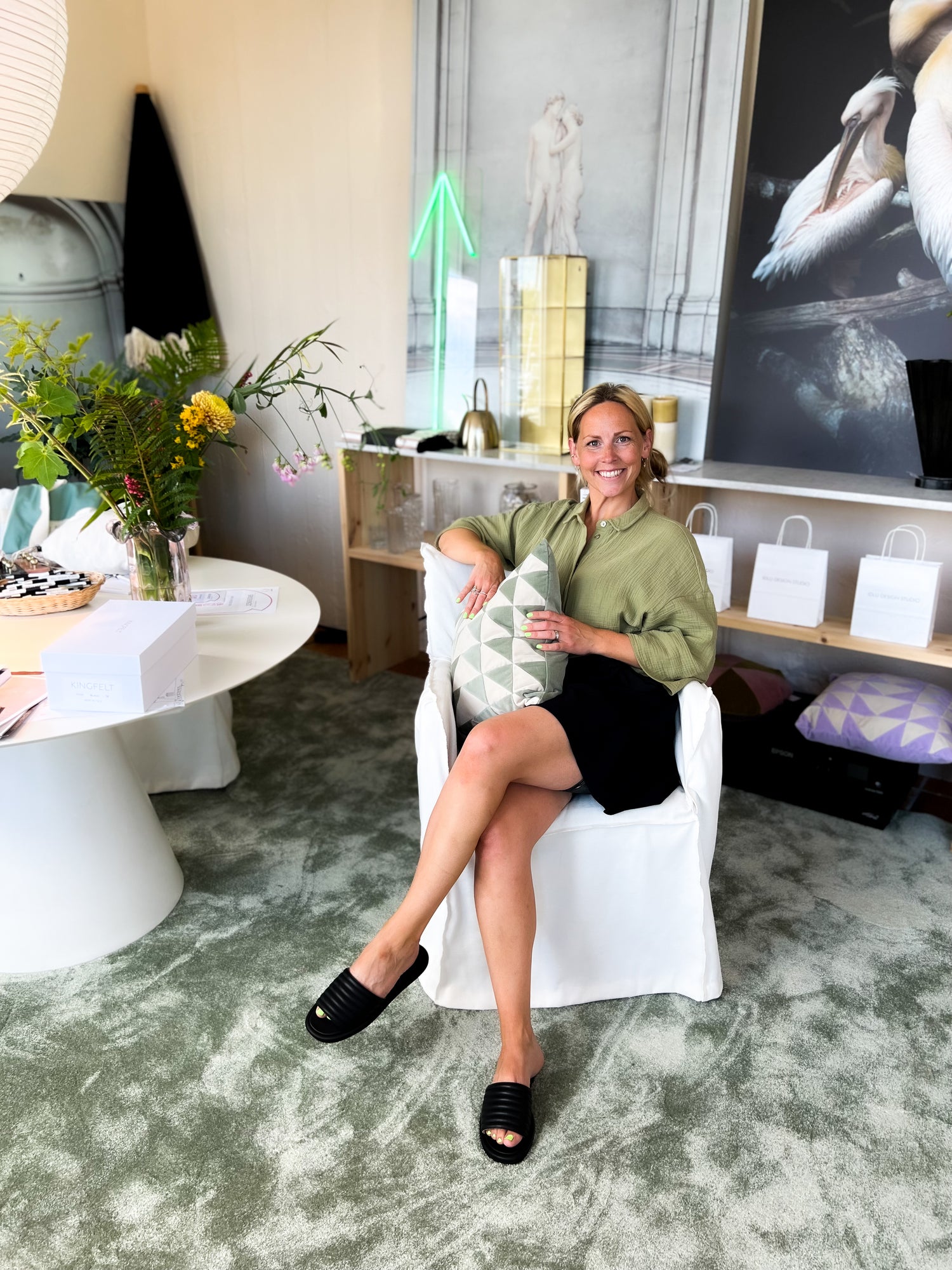 Svarta slip-in sandaler med en mjuk vadderad vågig ovandel med innerfoder av getskinn. Kreatören Ida sitter på en vit stol i design studion i Västervik.
