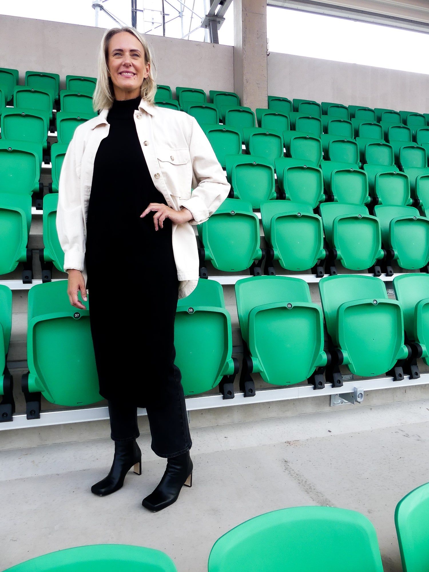 Personliga tränaren Lillemor står inne vid en stadium med gröna stolar i bakgrunden och grå cement. Hon bär KINGFELT FOOTWEARs svarta skinnstövletter av nappaskinn. Designade i Sverige och tillverkade i Italien.