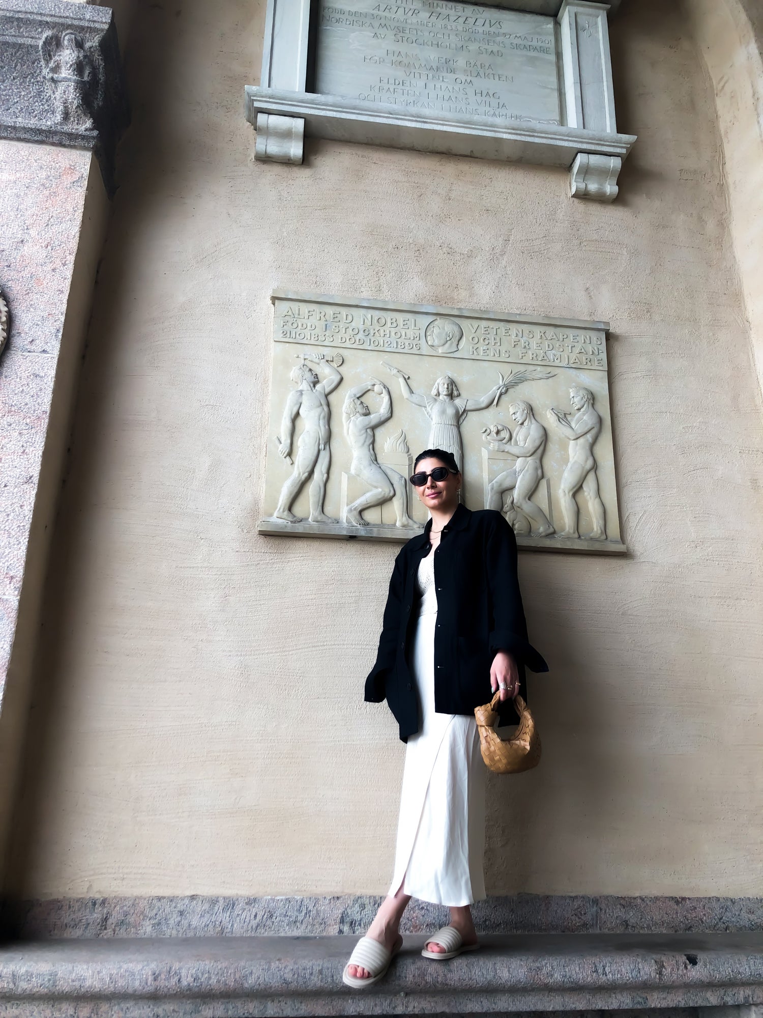 Kvinna står med beige kjol och svart kavaj vid Stockholm stadshus mot en vacker vägg. Hon bär KINGFELT FOOTWEARs beige slip-in sandaler Coco. Handgjorda och tillverkade i Italien.