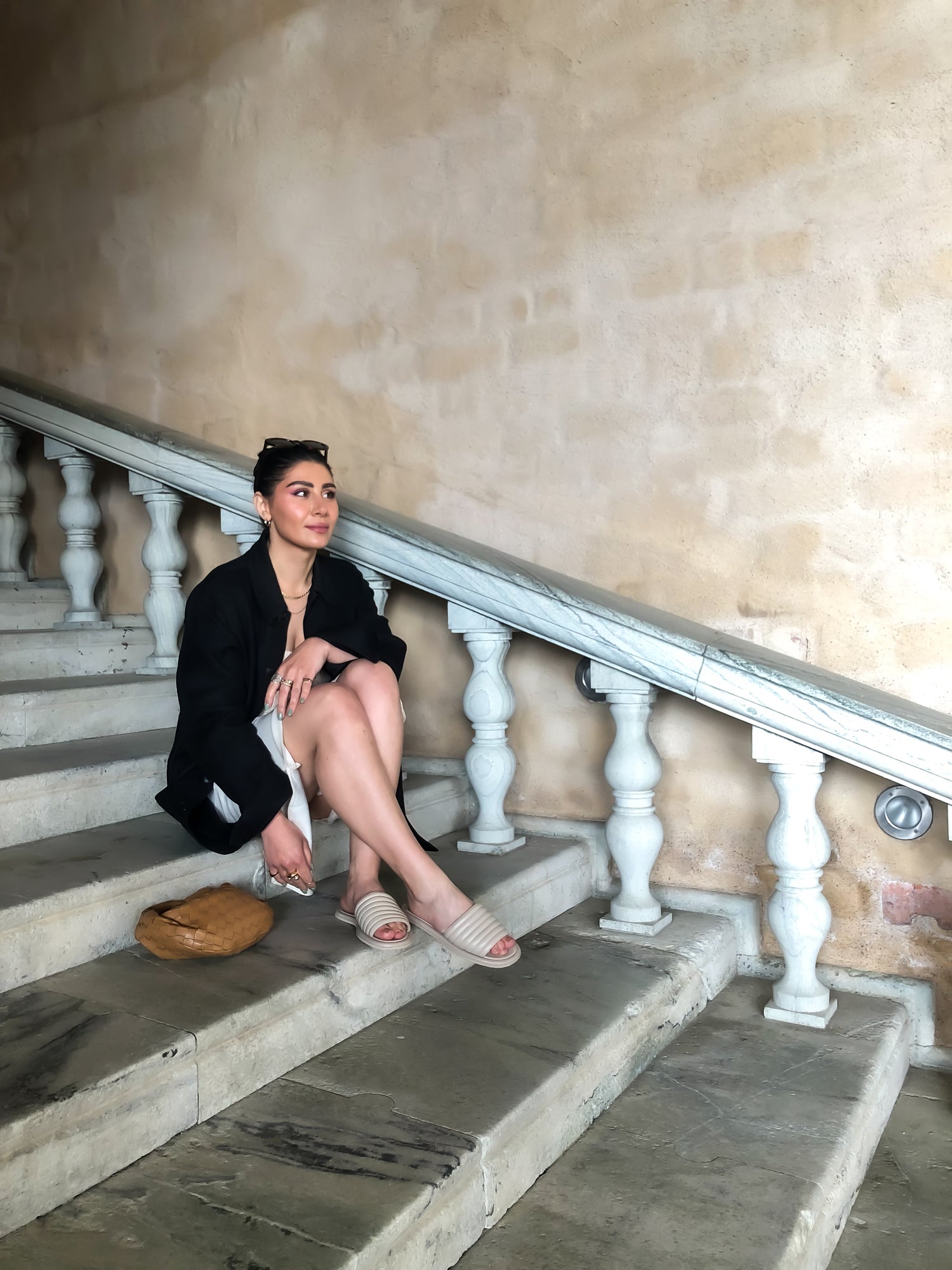 Gohar sitter på en fantastisk marmortrapp vid Stockholms stadshus med en ljusgul kalkvägg i bakgrunden.. Hon bär KINGFELT FOOTWEARs beige slip-in sandaler. Hållbart handgjordsa i Italien av hantverkare.