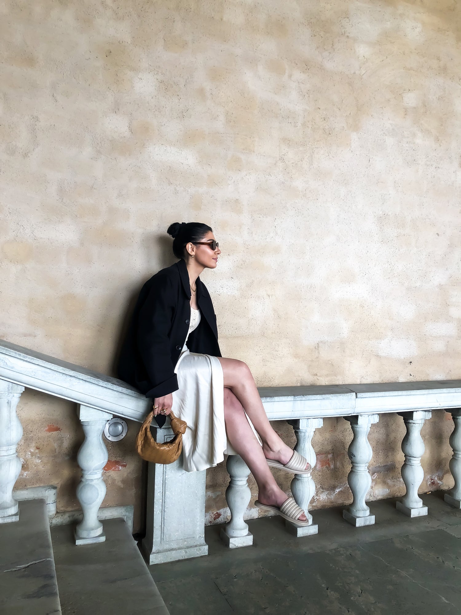 Ängelinvesteraren Gohar sitter likt en ballerina på ett trappräcke vid Stockholms stadshus för en plåtning med KINGFELT FOOTWEAR. Hon bär beige slip-in sandaler av mjukt nappaskinn.