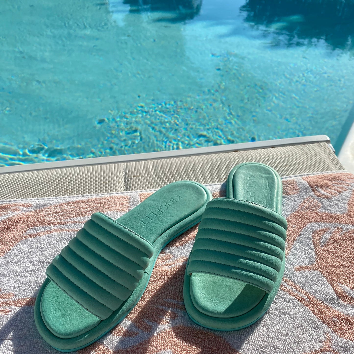 Mintgröna slip-in sandaler i mjukt kalvskinn ligger vid en pool på en vit ljusrosa handduk.Handgjorda och tillverkade i Italien.