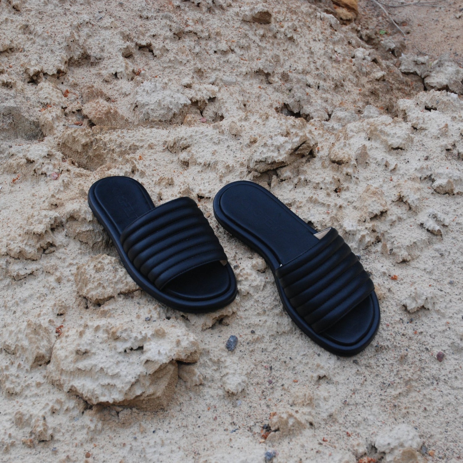 Style coco i svart är en slip-in sandal i kalvskinn. Handgjorda och tillverkade i Italien. Skorna ligger här på en sandstrand.
