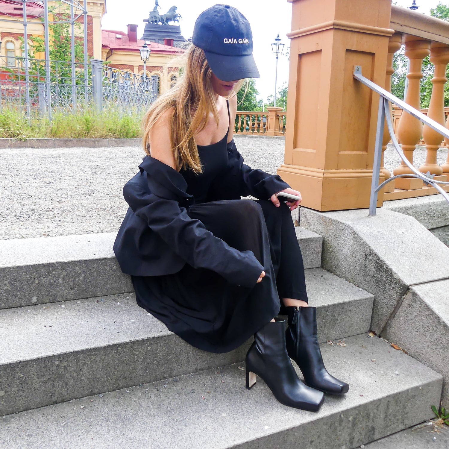 Hållbart producerade skinnstövletter i svart napaskinn. Tillverkade i Italien. Kvinna sitter på stentrapp vid cirkus i Stockholm med Kingfelt footwears square toe stövletter på sig. Skinnkläddklack med dold dragkedja.