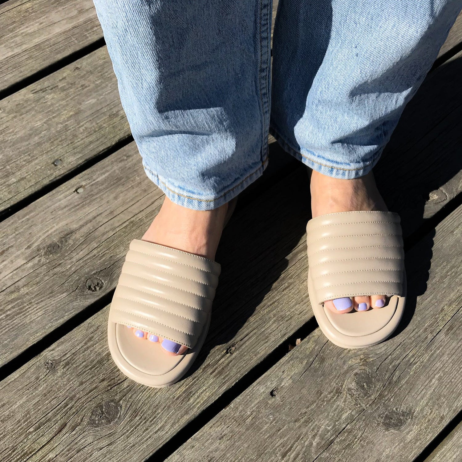 Beige slip-in sandal i nappaskinn. Öppen tå och lilla tånaglar i solen. Ljusa jeans på en träbrygga. Kingfelt footwear sandal. 