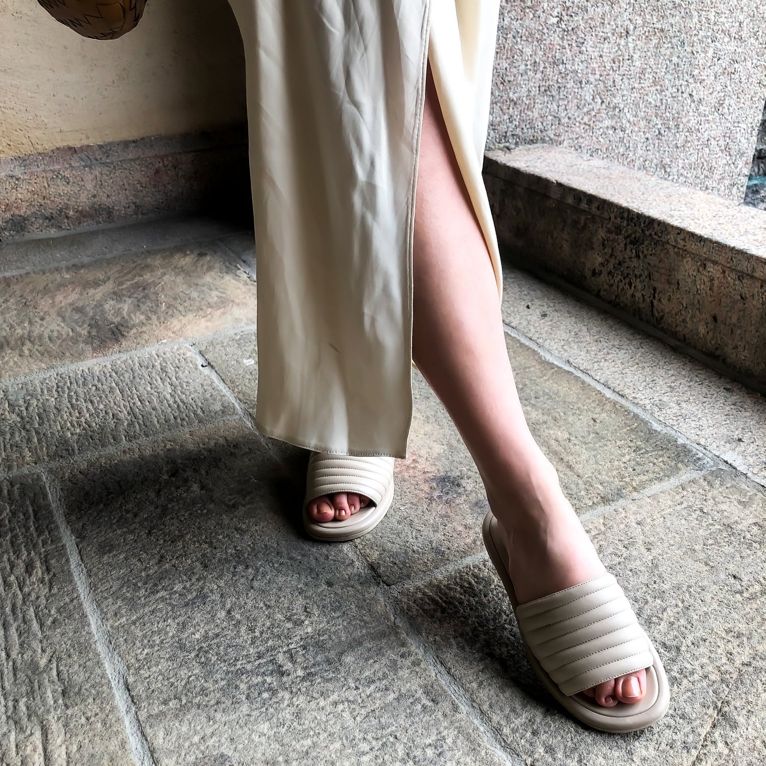Beige slip-in sandal av mjukt nappaskinn, tillverkade i Italien. Öppen tå sandal med vaddrad ovandel och innerfoder av getskinn. Pointad höger fot med beiged rosa nagalelack och en vit kjol med slitz vid stadshuset i Stockholm.